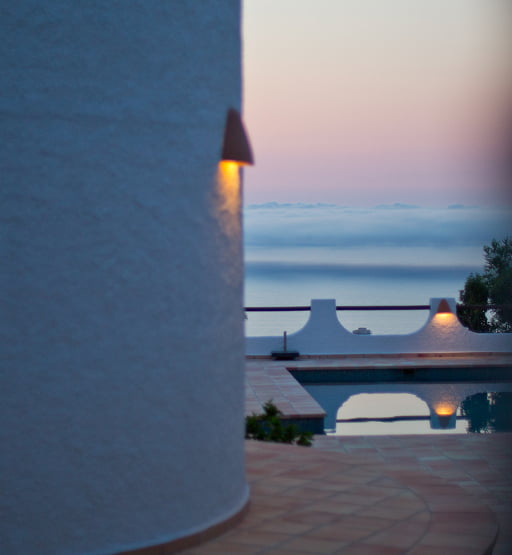 Pared blanca con apliques encendidos, piscina y vistas al mar al atardecer.