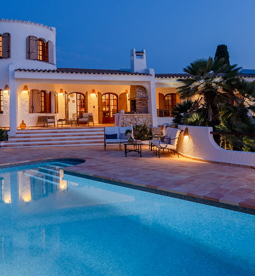 Vista nocturna de una villa blanca con iluminación cálida y piscina.